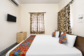 A hotel room with a bed, a TV mounted on the wall, a wooden nightstand, and a window with patterned curtains.