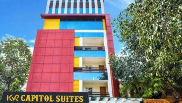 Un hotel de varios pisos con una fachada destacada de color rojo y amarillo, una entrada visible y un letrero que dice 'KR CAPITOL SUITES'.