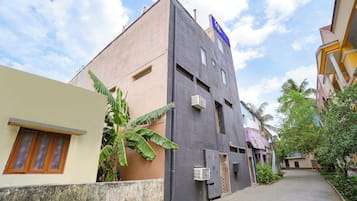 A narrow street with buildings, including a two-story structure with a blue sign, a small window, and an air conditioning unit.