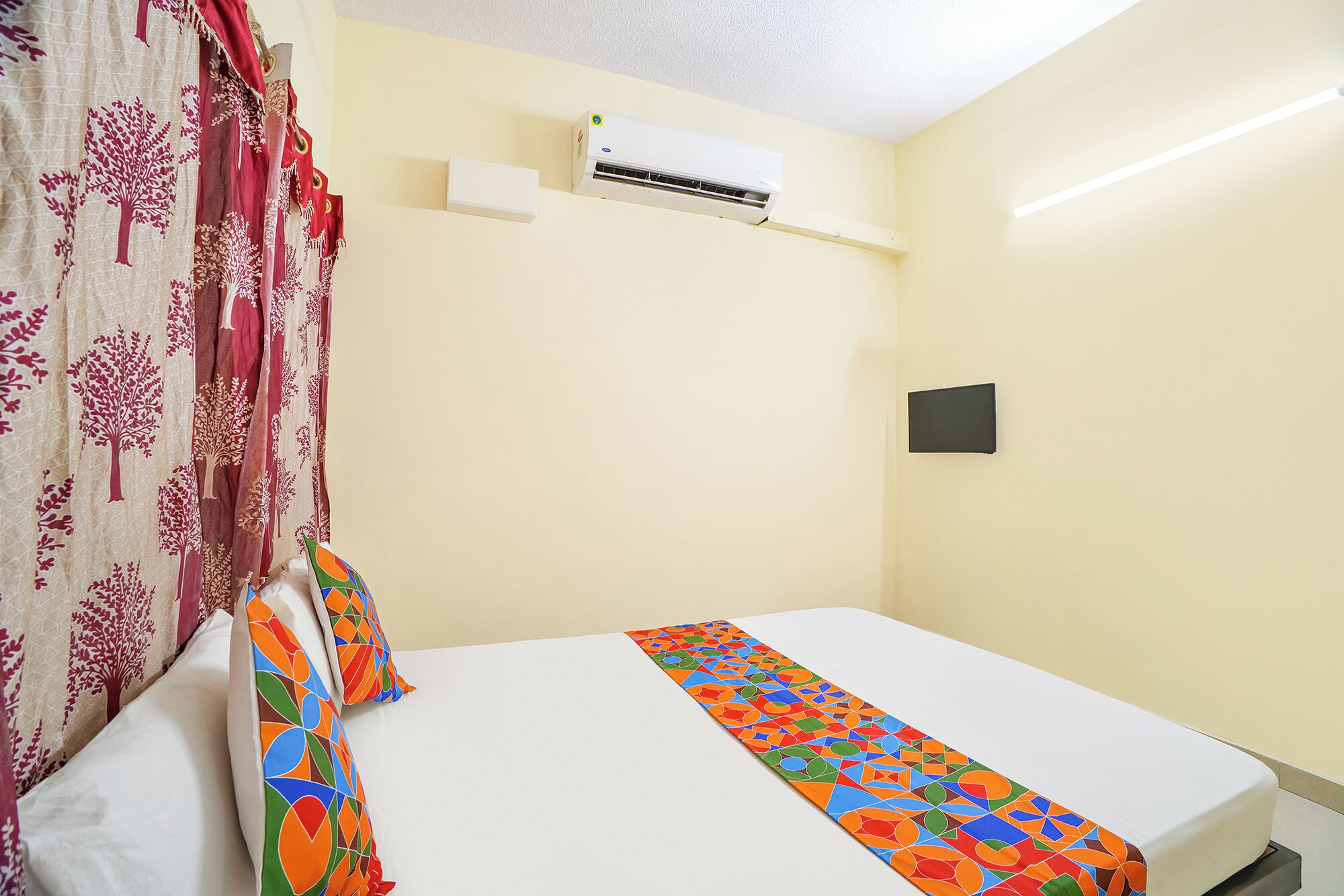 A hotel room with a bed, a window with curtains, an air conditioning unit, and a wall-mounted light switch.