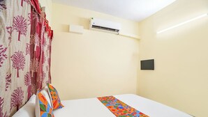 A hotel room with a bed, a window with curtains, an air conditioning unit, and a wall-mounted light switch.