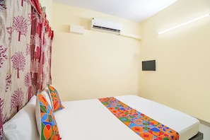 A hotel room with a bed, a window with curtains, an air conditioning unit, and a wall-mounted light switch.