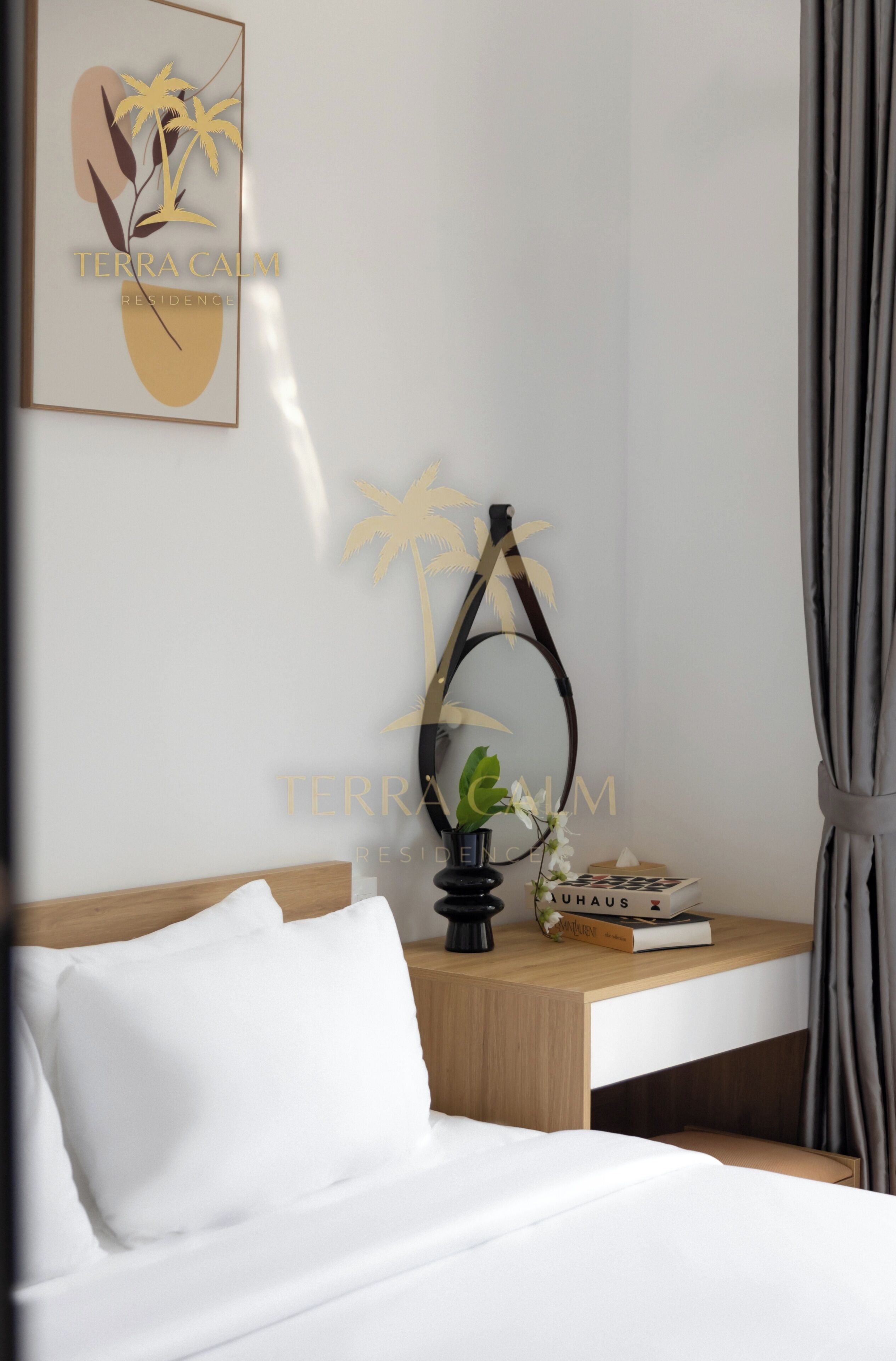 Una cama bien hecha con sábanas blancas, una mesita de noche de madera con un libro y una planta pequeña, y una obra de arte colgada en la pared que representa palmeras y una forma abstracta.