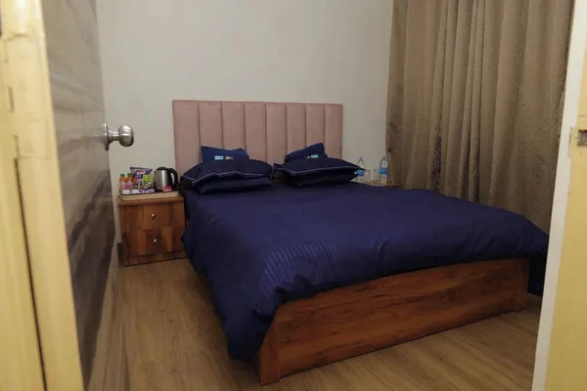 Una habitación de hotel con piso de madera, una cama con cabecera, una mesita de noche con hervidor y una cortina.