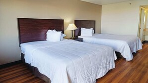 A hotel room with two beds, wooden flooring, a nightstand with a lamp, and a door leading to another room.