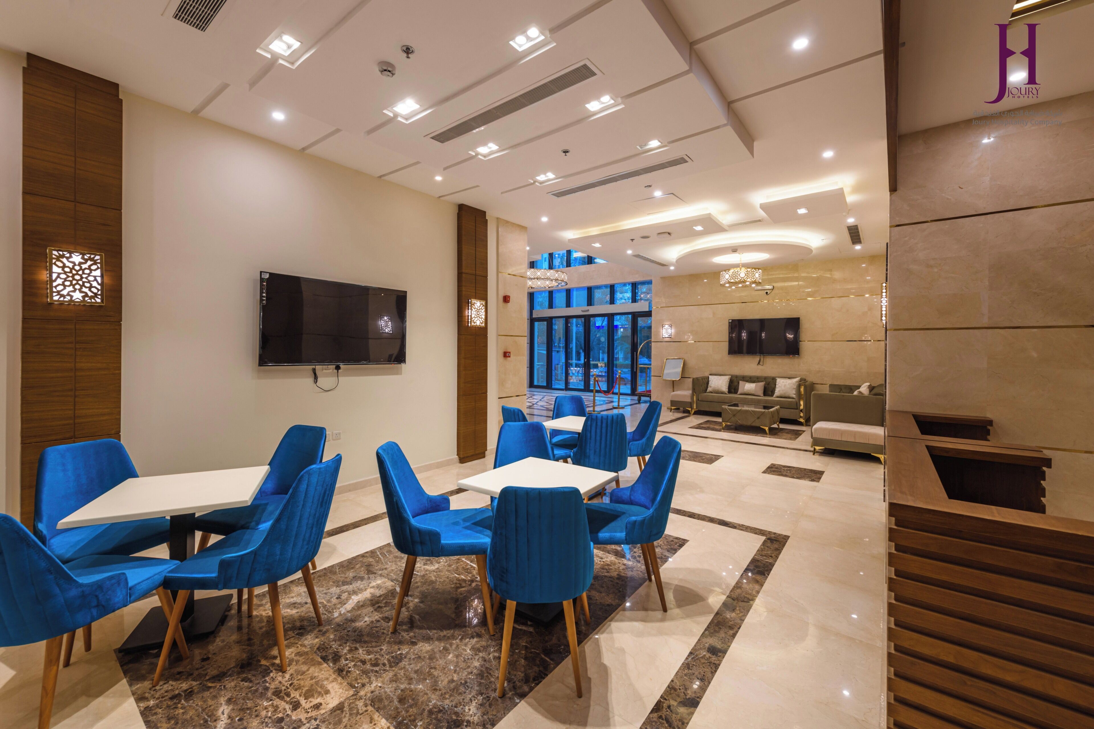 Vestíbulo de hotel con diseño moderno, que incluye una zona de estar con sillas azules y una mesa blanca, un televisor de pantalla plana montado en la pared y un mostrador de recepción al fondo.