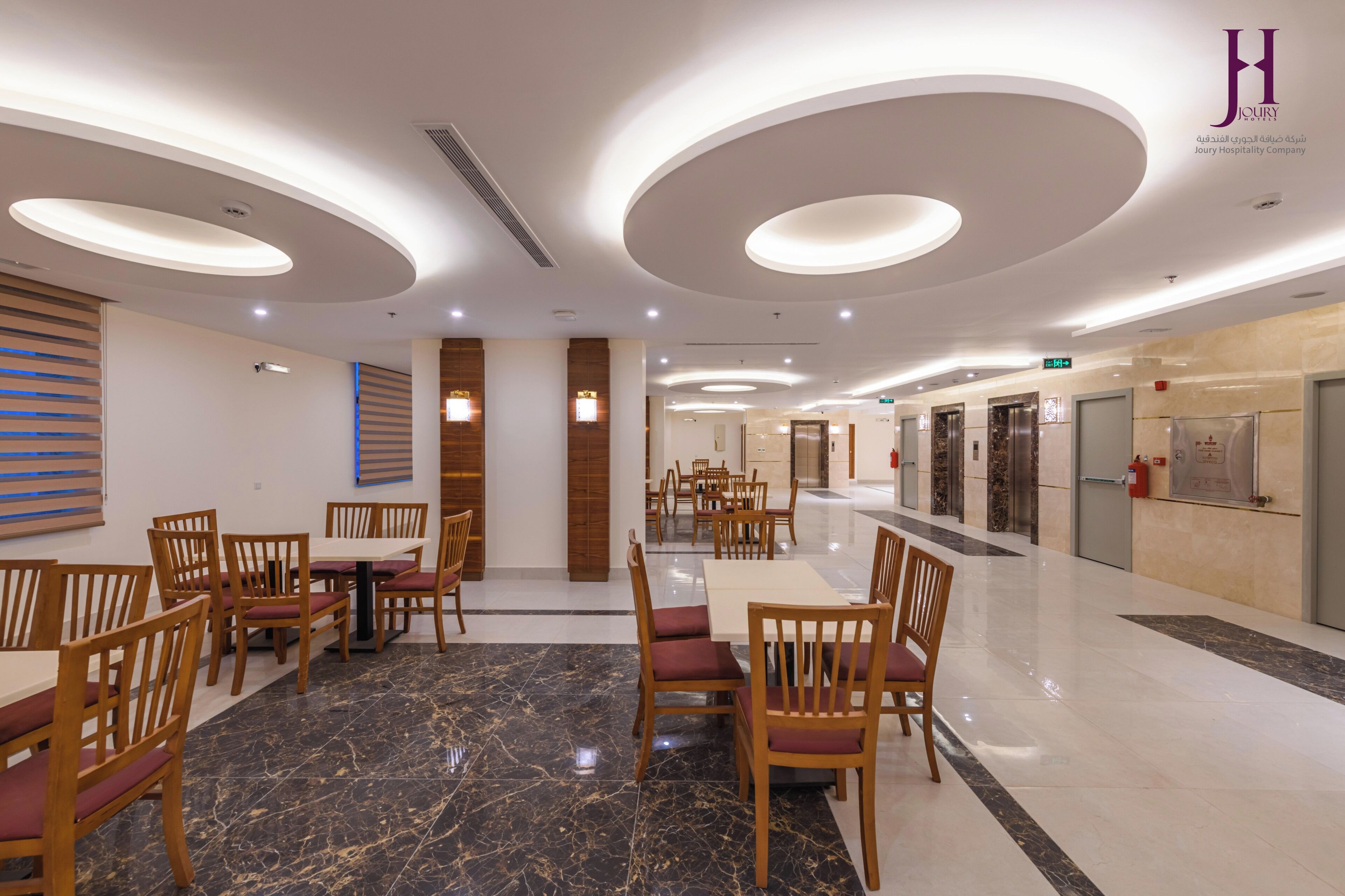Un hall de hotel con diseño moderno, que presenta un área de estar central con mesas y sillas, un suelo de mármol y un techo con lámparas circulares.