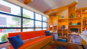 A room with a wooden bench, a desk with a computer, shelves with various items, and a window with a view of the outside.