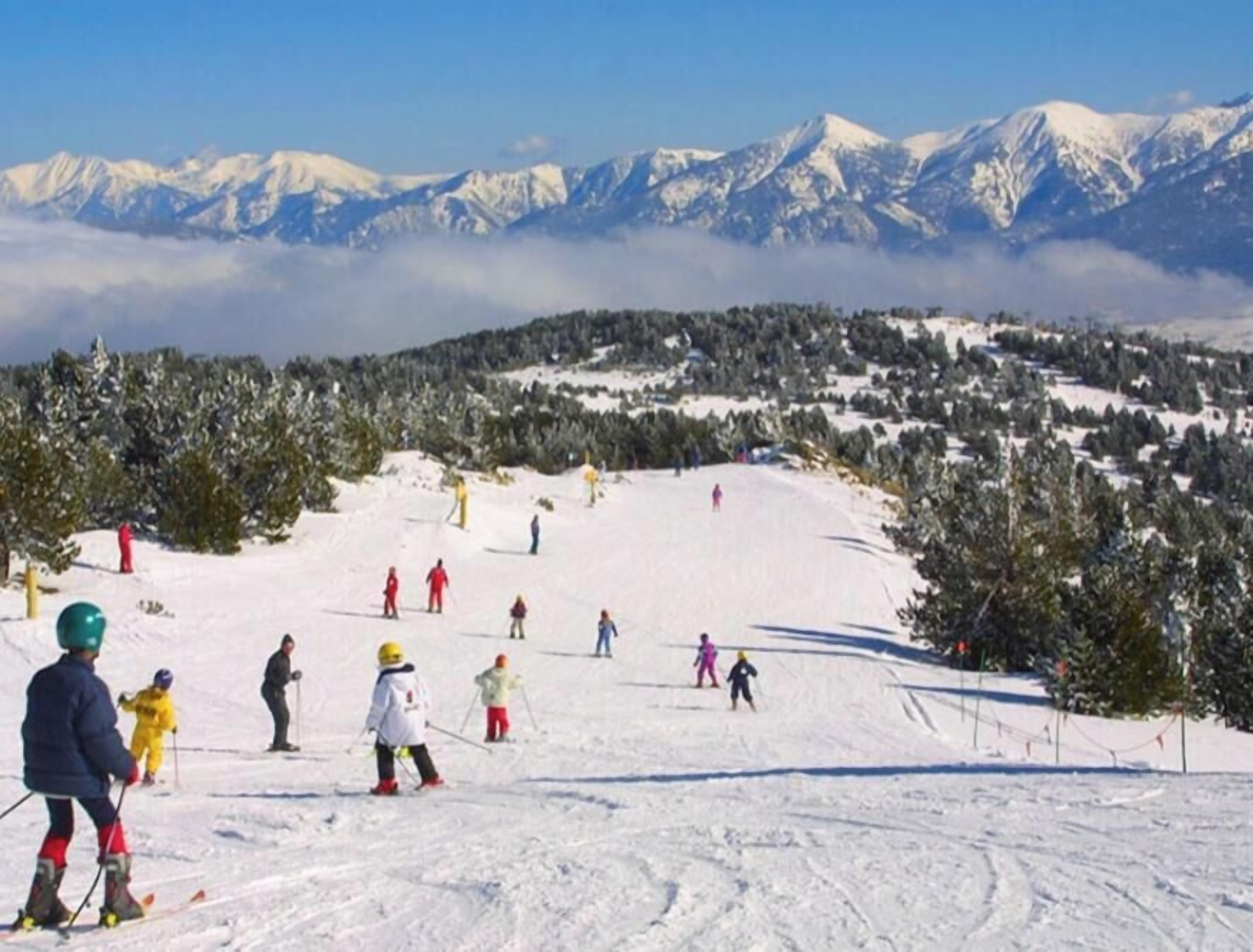 Una pista de esquí con esquiadores y snowboarders, rodeada de montañas y árboles cubiertos de nieve.
