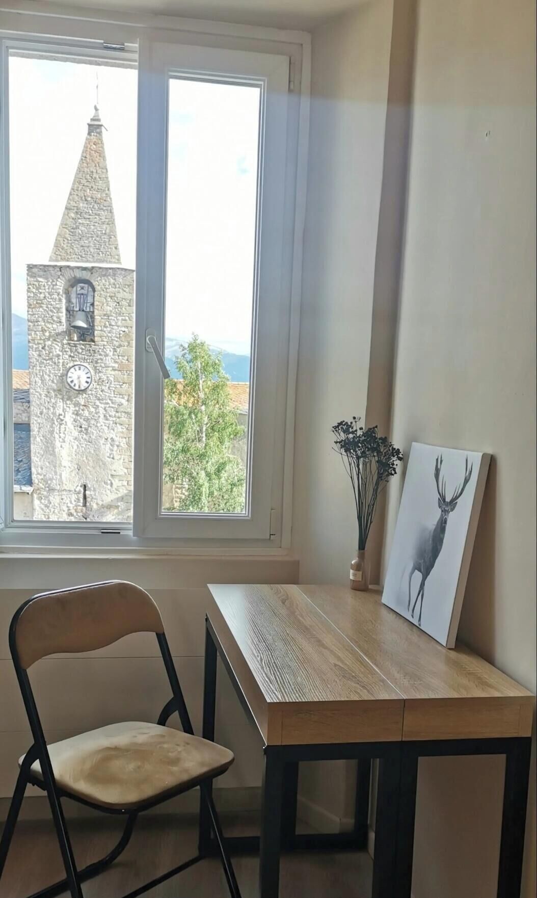 Een kamer met een houten tafel, een stoel, een schilderij van een hert, een raam met uitzicht op een stenen toren en een vaas met bloemen.