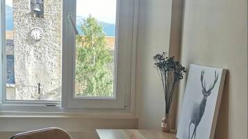 Een kamer met een houten tafel, een stoel, een schilderij van een hert, een raam met uitzicht op een stenen toren en een vaas met bloemen.