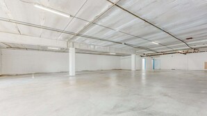 Un espacio amplio y vacío de estilo industrial, con suelos de hormigón y paredes blancas, y tuberías del techo a la vista, además de algunas puertas.