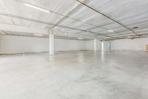 A large, empty industrial-style room with concrete floors and white walls, featuring exposed ceiling pipes and a few doors.