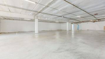 Een ruime, lege industriële ruimte met betonnen vloeren en witte muren, voorzien van zichtbare leidingen in het plafond en enkele deuren.