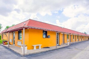Un motel con techo rojo y paredes exteriores amarillas, que presenta una hilera de habitaciones y un pequeño espacio de estar al aire libre.
