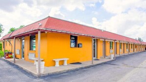 Un motel con techo rojo y paredes exteriores amarillas, que presenta una hilera de habitaciones y un pequeño espacio de estar al aire libre.