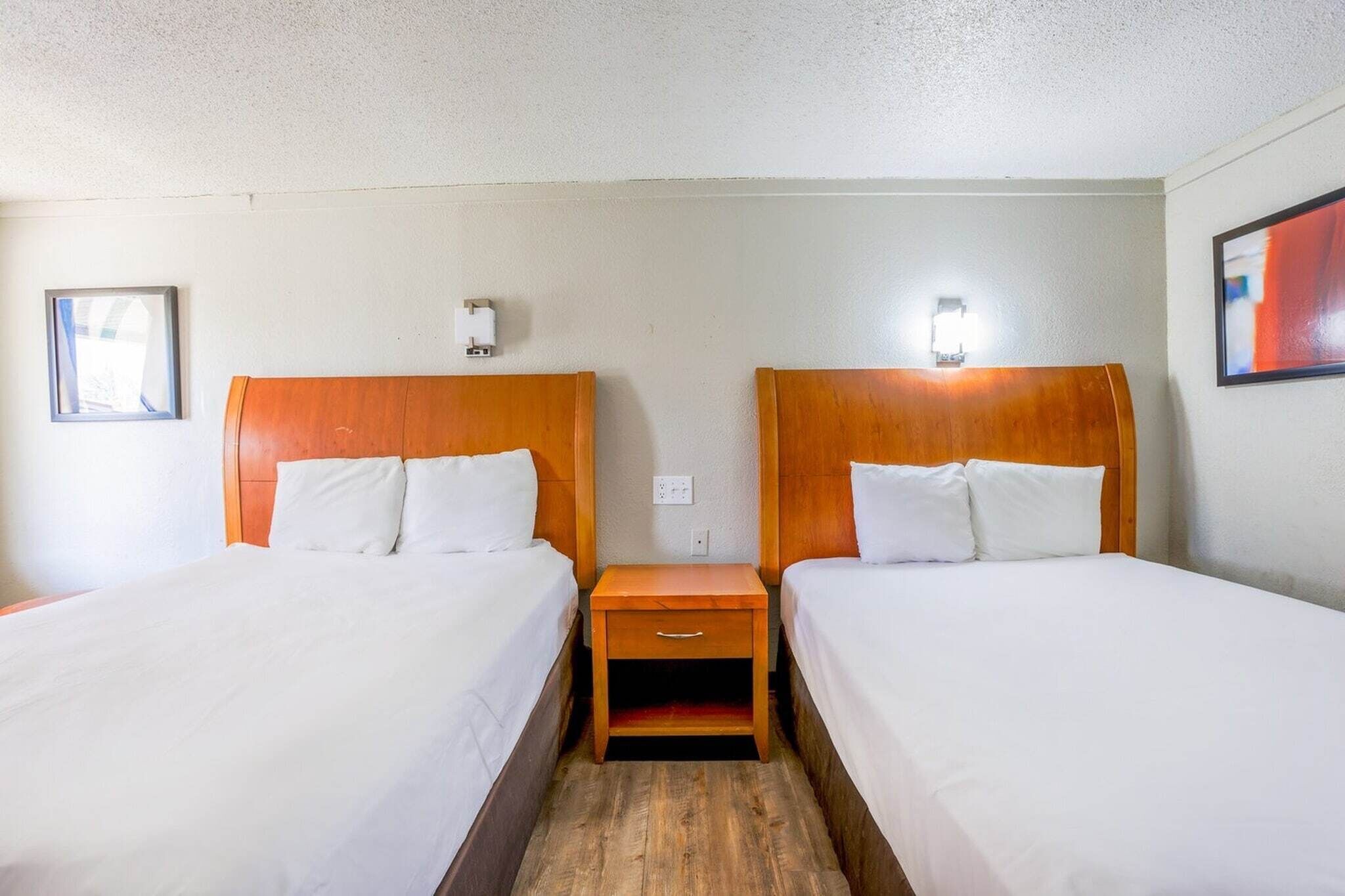 A hotel room with two beds, a wooden nightstand, a wall socket, and a framed picture.