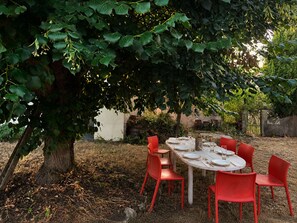 An outdoor dining area with a table set for six, surrounded by red chairs, under a large tree.