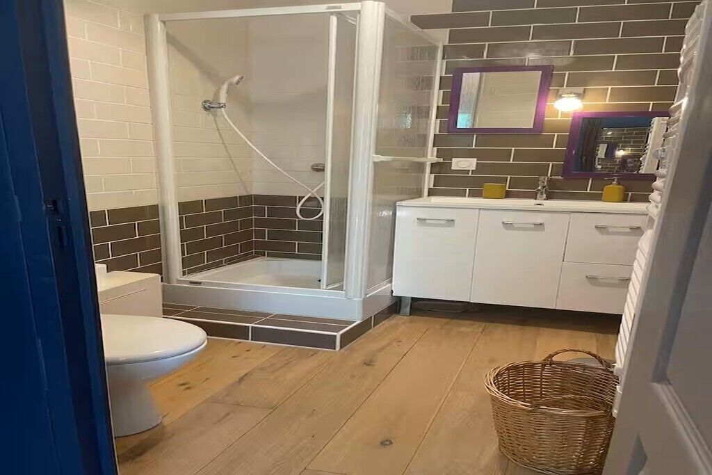 A bathroom with a glass-enclosed shower, a toilet, a sink with a mirror, and a basket on the floor.