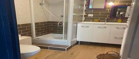 A bathroom with a glass-enclosed shower, a toilet, a sink with a mirror, and a basket on the floor.
