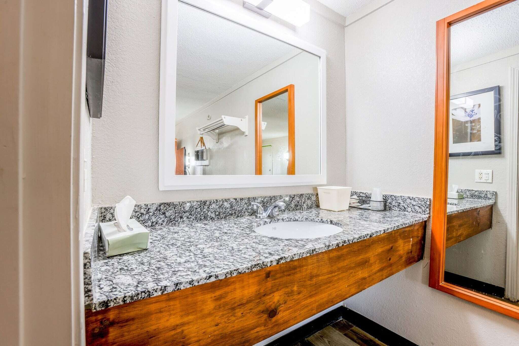 A bathroom with a granite countertop, a white sink, a wooden vanity, a large mirror, and a framed picture on the wall.