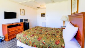 A hotel room with a bed, a wooden dresser, a television, a desk with a lamp, and framed pictures on the walls.