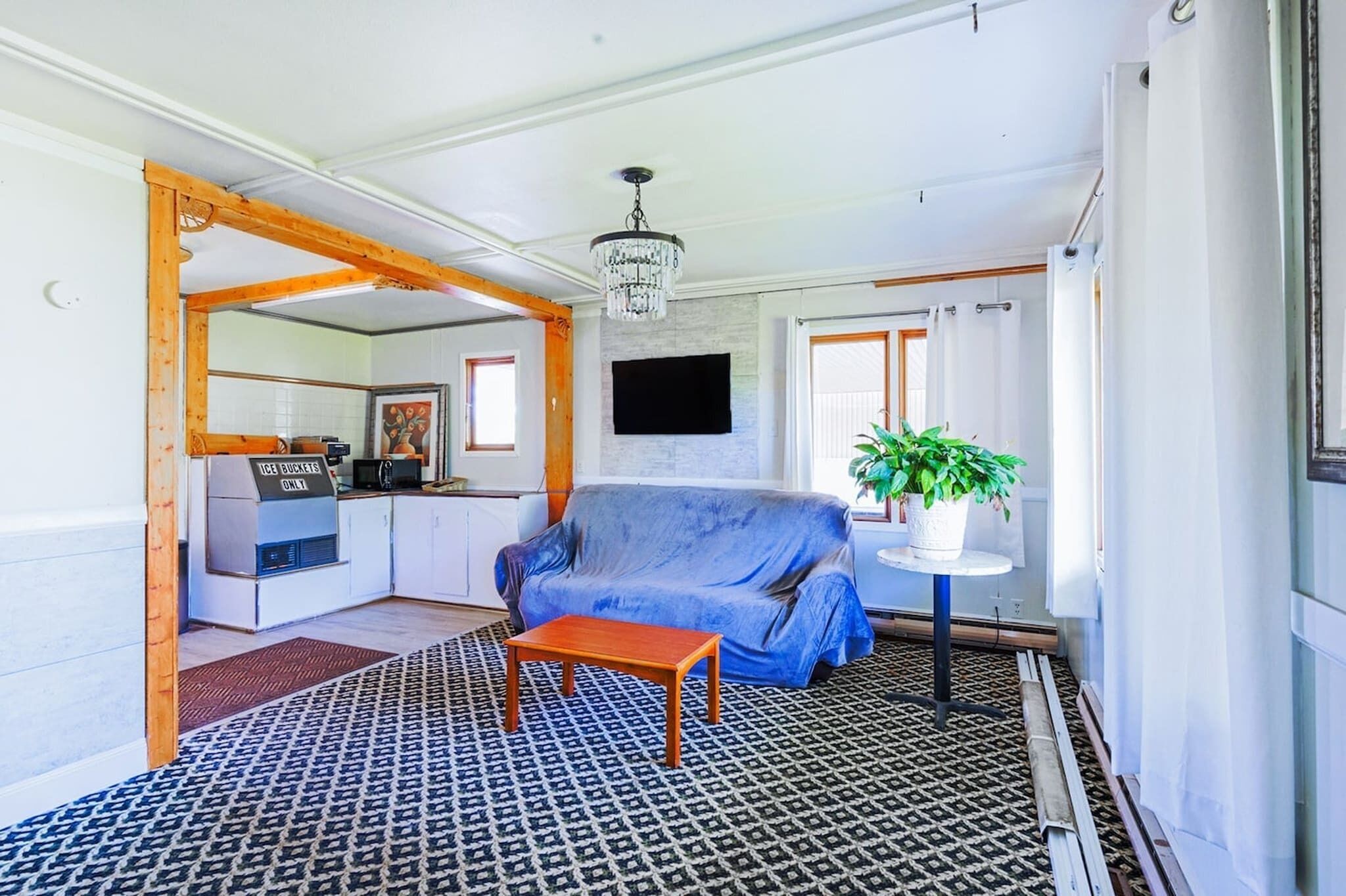 Ein Wohnzimmer mit blauem Sofa, einem Holztisch, einem gemusterten Teppich und einer Chandelier. Im Küchenbereich ist ein Schild mit der Aufschrift 'ICE BUCKETS ONLY'.