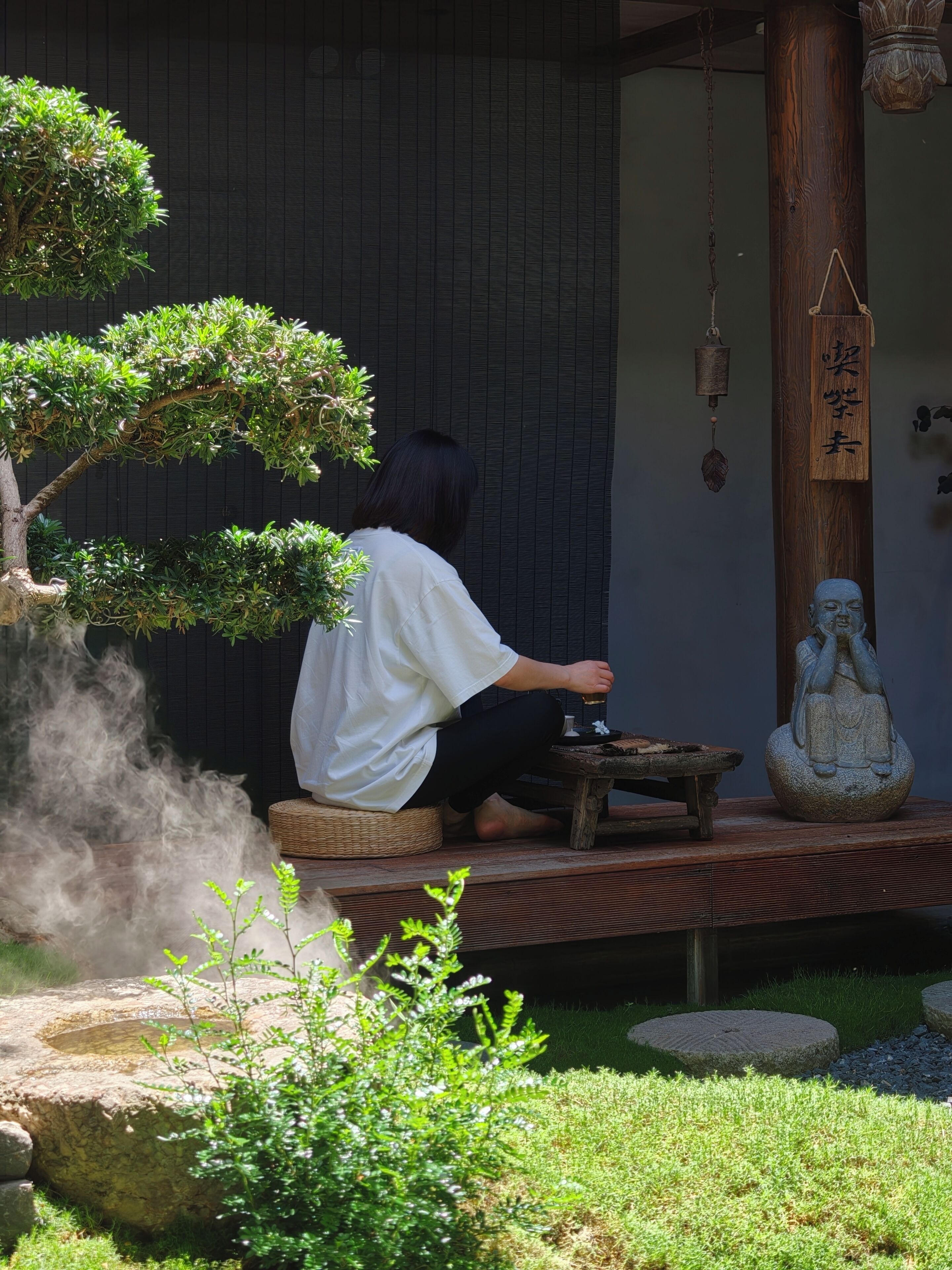 A person sitting on a bench in a garden with a bonsai tree, a stone statue, and a wooden structure with Chinese characters.