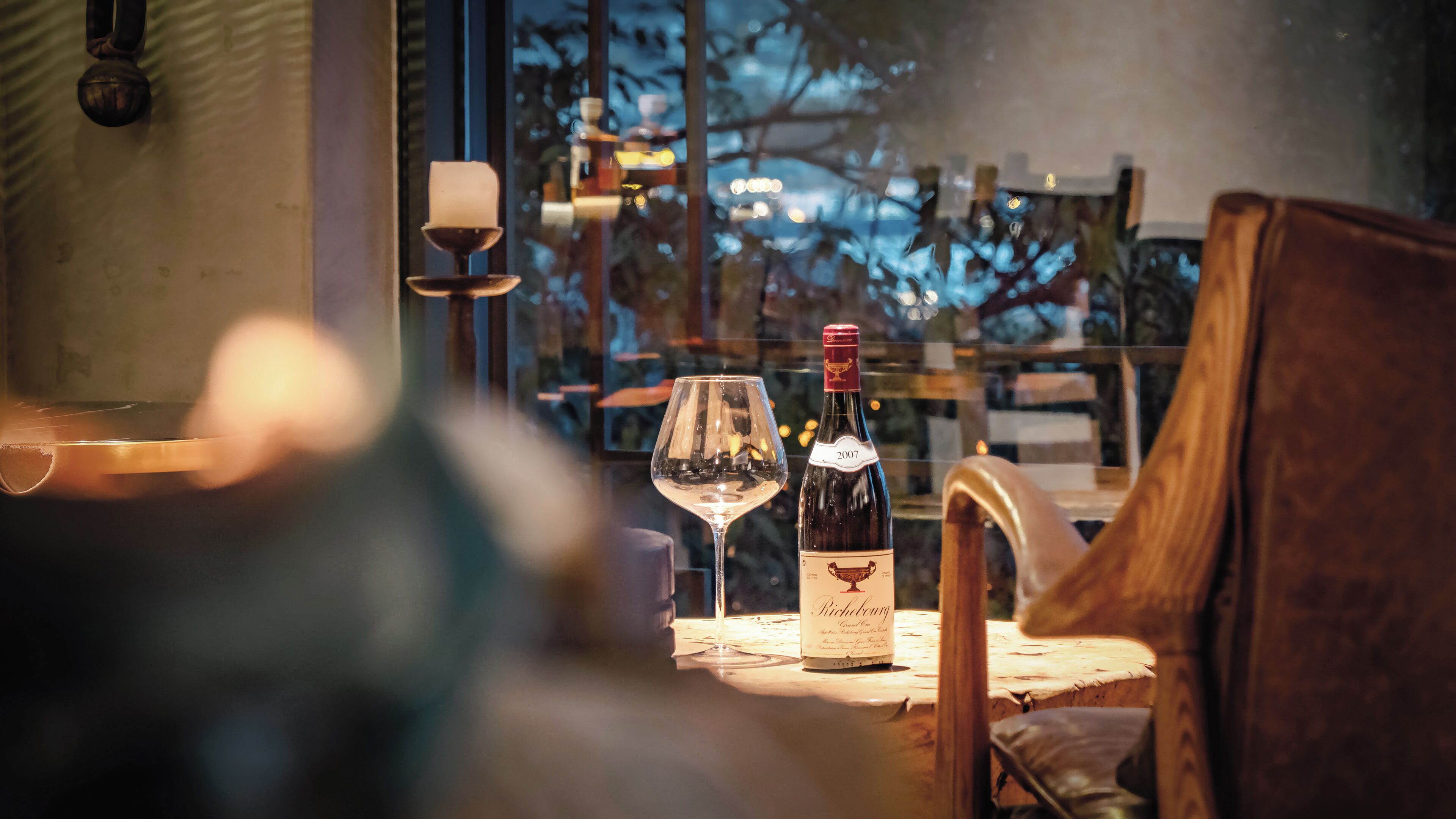 Una mesa con dos botellas de vino y una copa, una vela y vistas de árboles a través de una ventana.