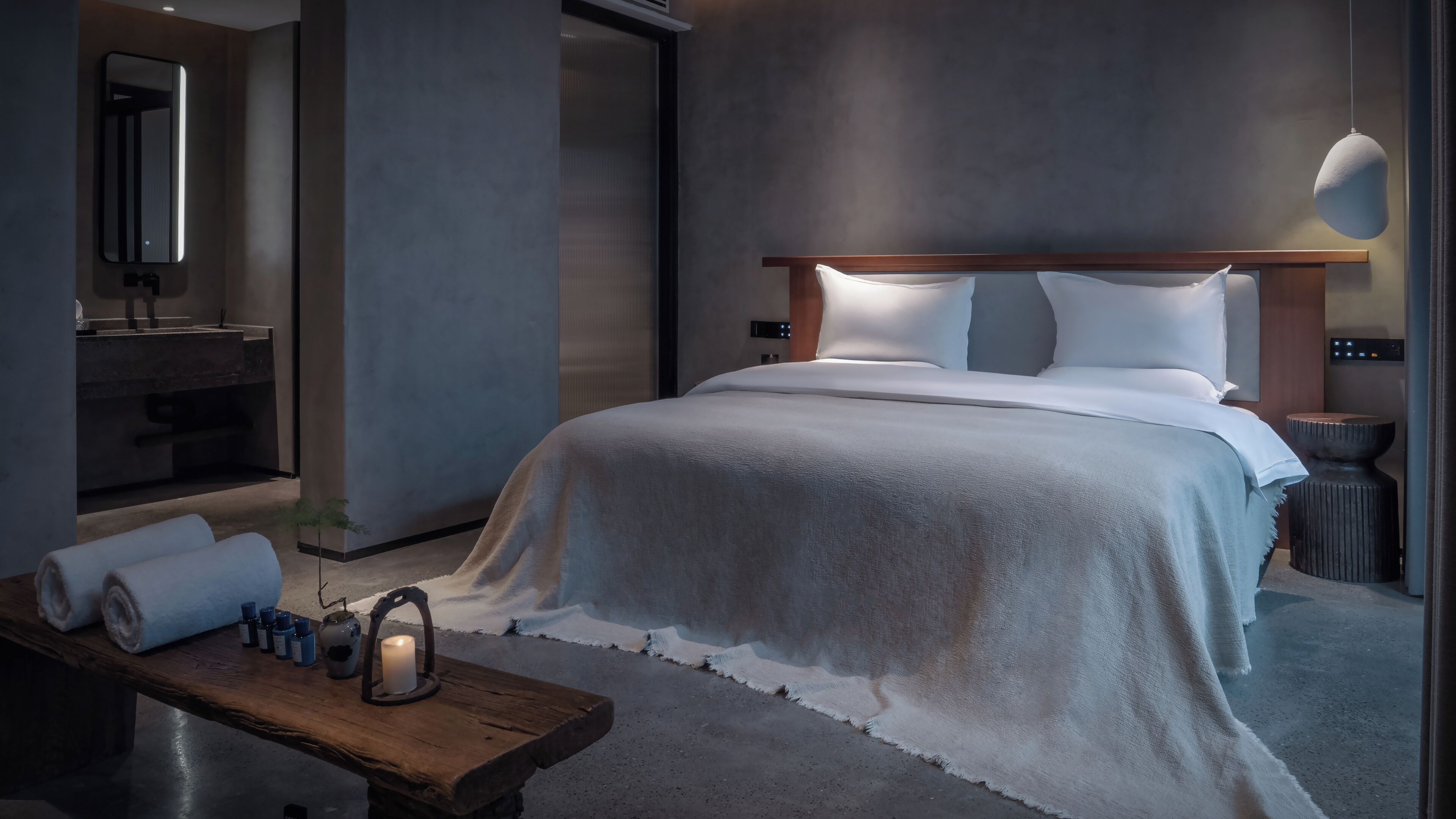 A modern hotel room with a large bed, a wooden nightstand, a mirror, and a bathroom with a sink and mirror.