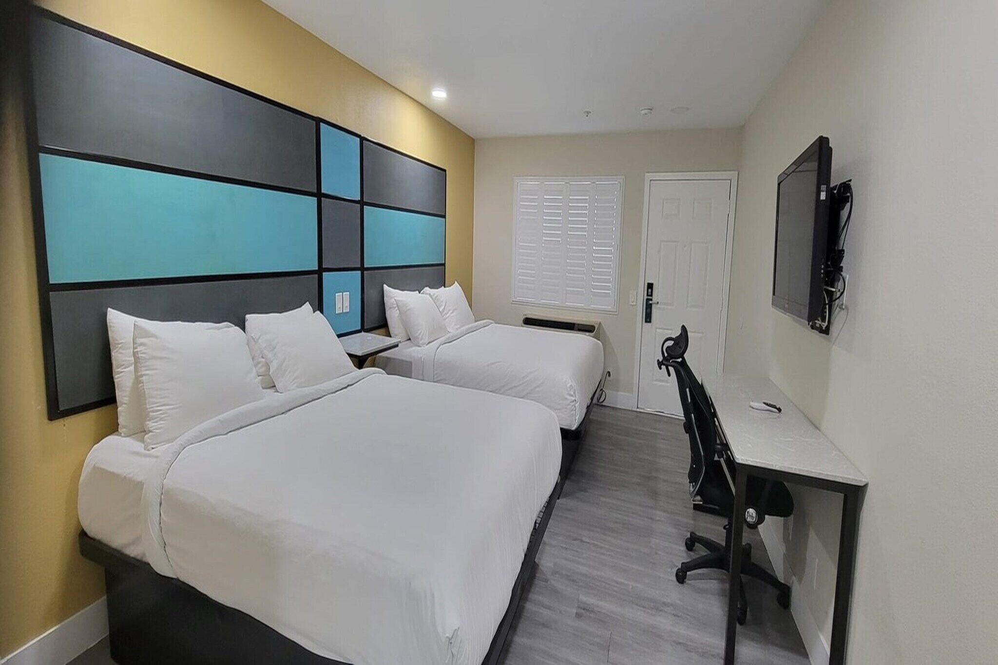 A hotel room with two beds, a desk, a chair, a television, and a door.