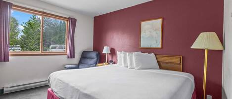 Une chambre d’hôtel avec un grand lit, une chaise, un bureau, une lampe et une fenêtre avec des rideaux.