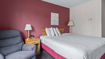 A hotel room with a bed, a chair, a nightstand, a lamp, and a painting on the wall.