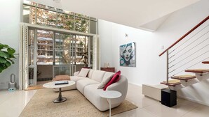 Ein modernes Wohnzimmer mit einem weißen Sofa, einem Couchtisch, einem kleinen Beistelltisch, einer Treppe mit einer Holzhandläufer und einem großen Fenster mit Blick auf Bäume und Gebäude.