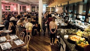 Ein belebtes Restaurant mit Holztischen und -stühlen, einem Barbereich mit Obstauslage und einer Küche im Hintergrund.