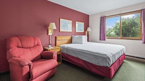 A hotel room with a bed, a red armchair, two bedside lamps, and a window with curtains.