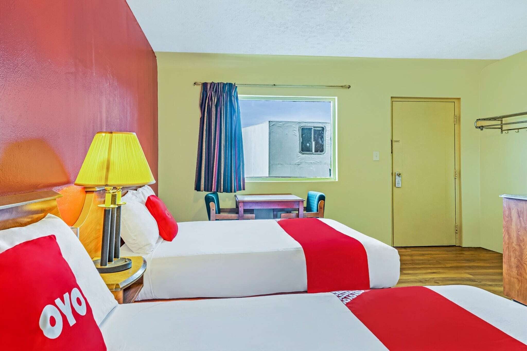 A hotel room with two beds, a table, chairs, a lamp, a window with curtains, and a door.