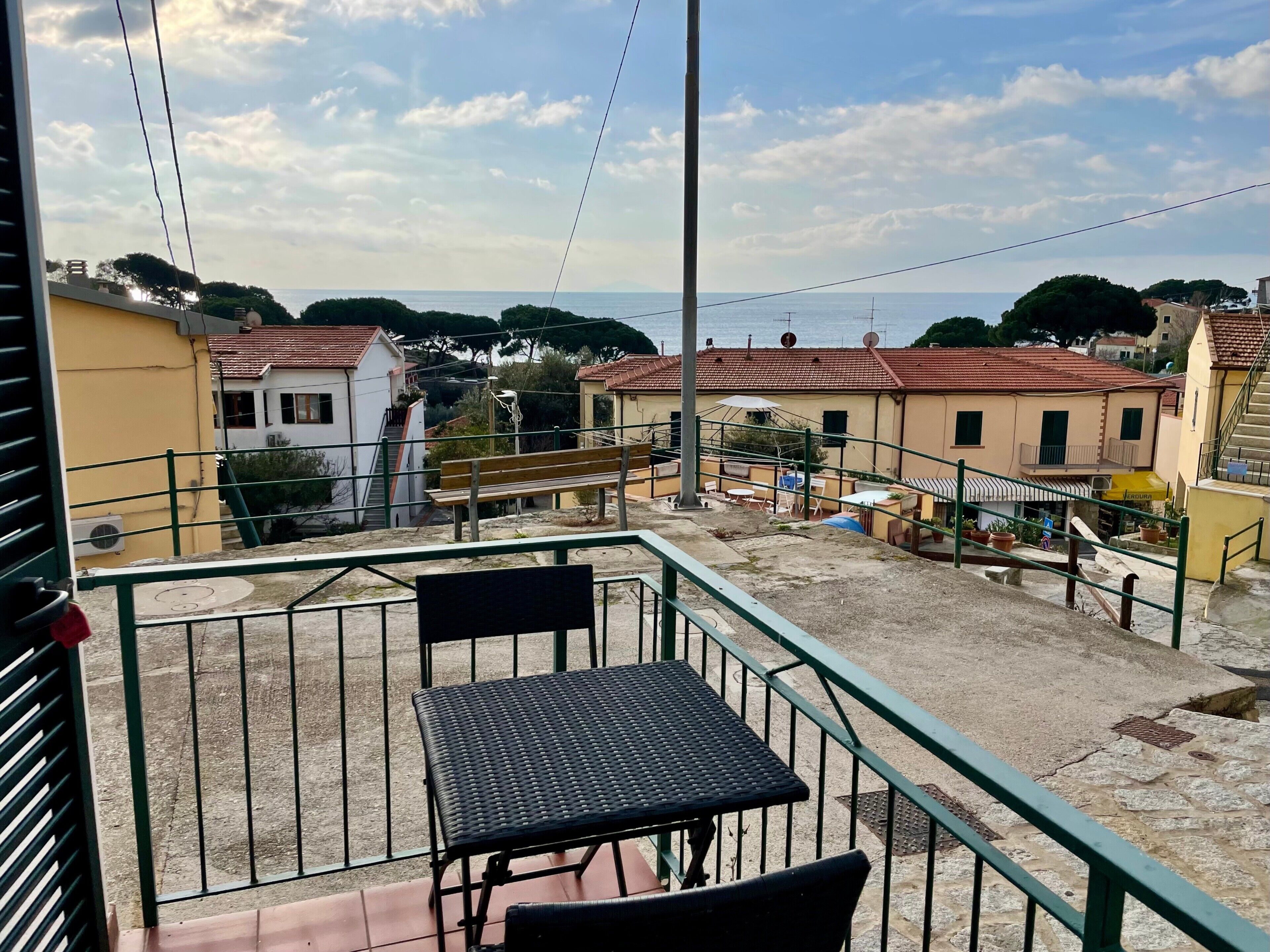 Un balcone con tavolo e sedie, affacciato su una zona residenziale con case e vista sul mare.
