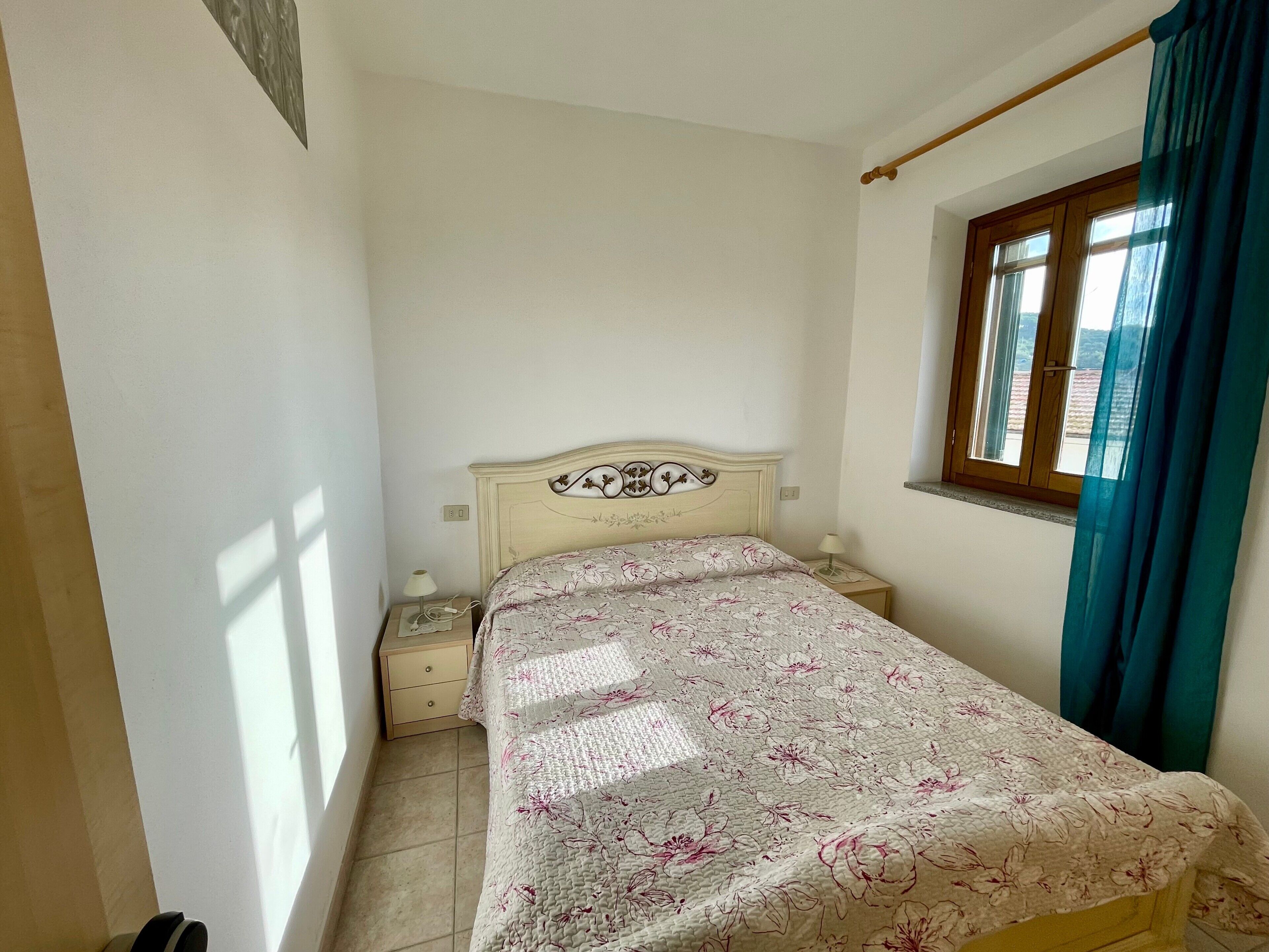 A bedroom with a bed, a nightstand, a window with curtains, and a wall-mounted light switch.