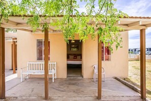 A covered outdoor seating area with a bench and chairs, a doorway leading to a room with a bed and a window, and a view of the surrounding area.