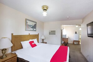 Une chambre d’hôtel avec un lit, une table de chevet, une lampe, un luminaire au plafond, un tableau au mur, une porte donnant sur une autre pièce et un téléviseur fixé au mur.