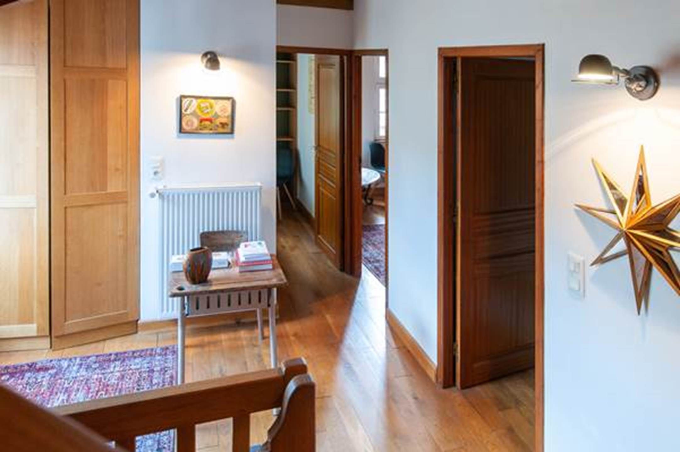 Un couloir avec des portes en bois, un radiateur, une table avec des livres et un élément de décoration murale en forme d’étoile.
