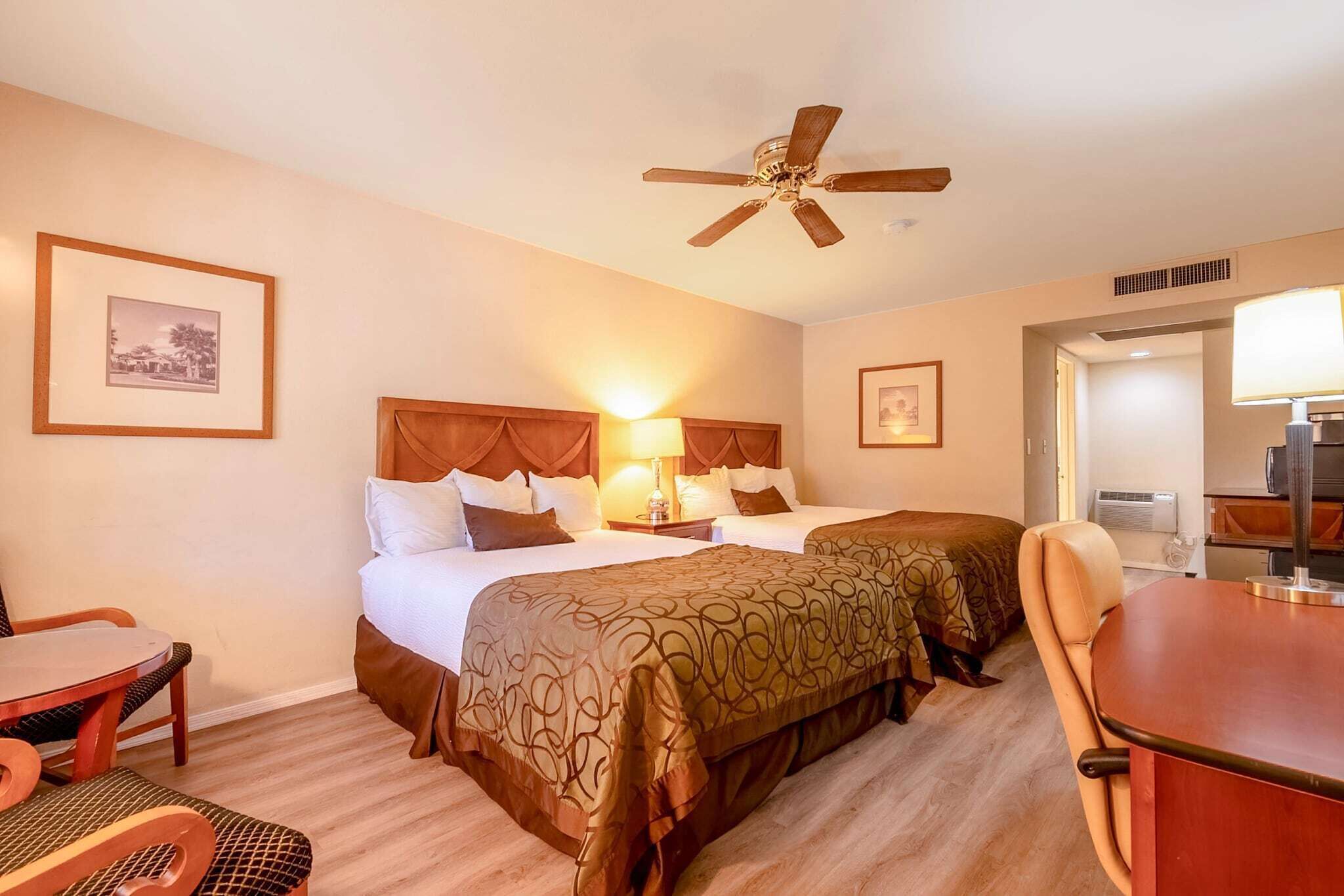 Een hotelkamer met twee bedden, een plafondventilator, een tafel met stoelen, en ingelijste foto’s aan de muur.