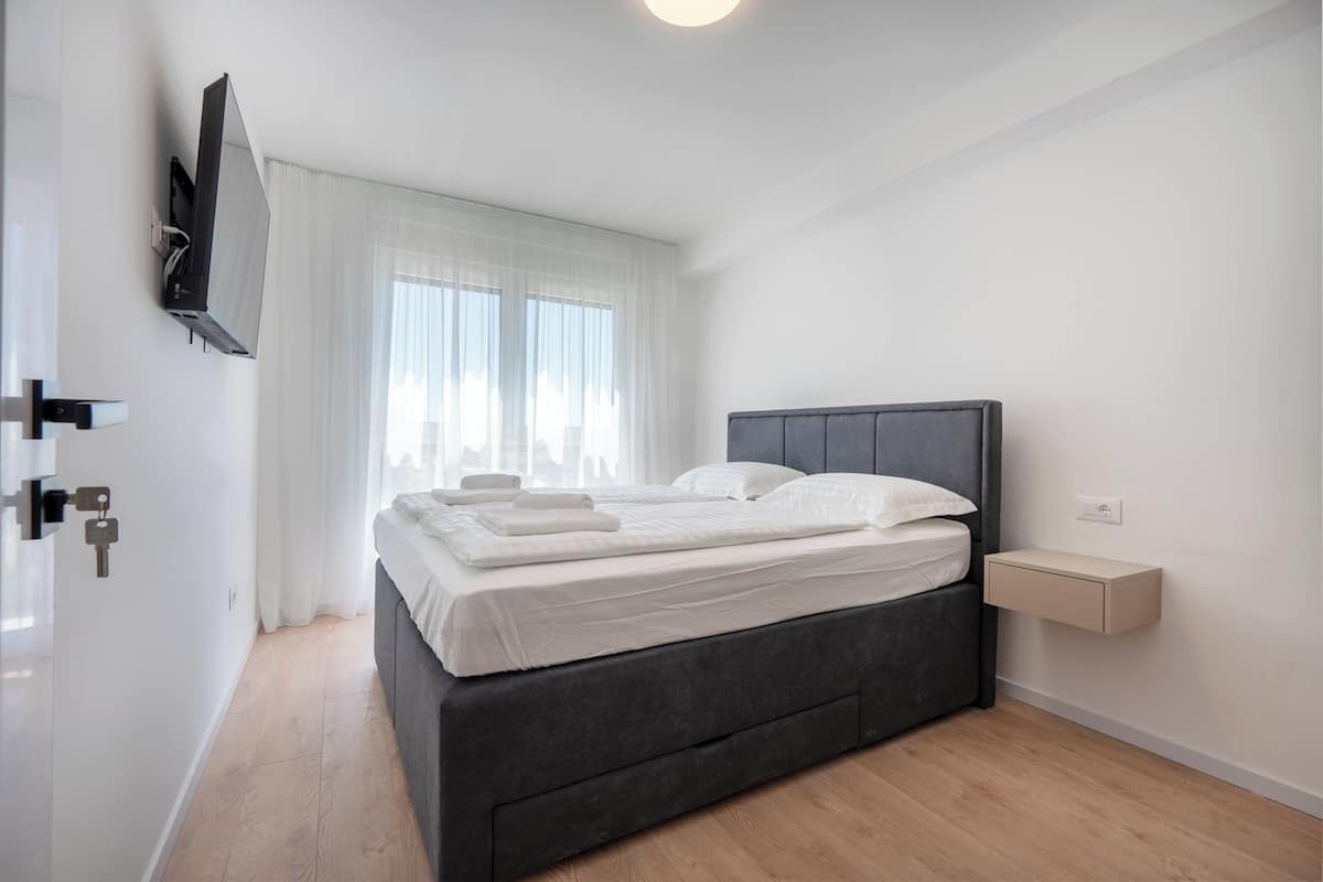 Une chambre à coucher moderne, avec un lit, une table de chevet, un téléviseur fixé au mur et une fenêtre ornée de rideaux légers.