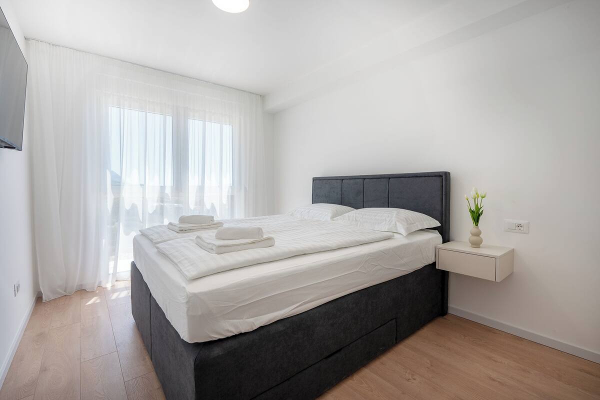 Une chambre à coucher moderne avec une tête de lit sombre, une table de chevet ornée d’un vase et une grande fenêtre avec des rideaux légers.