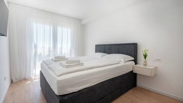 A modern bedroom with a dark headboard, a bedside table with a vase, and a large window with sheer curtains.