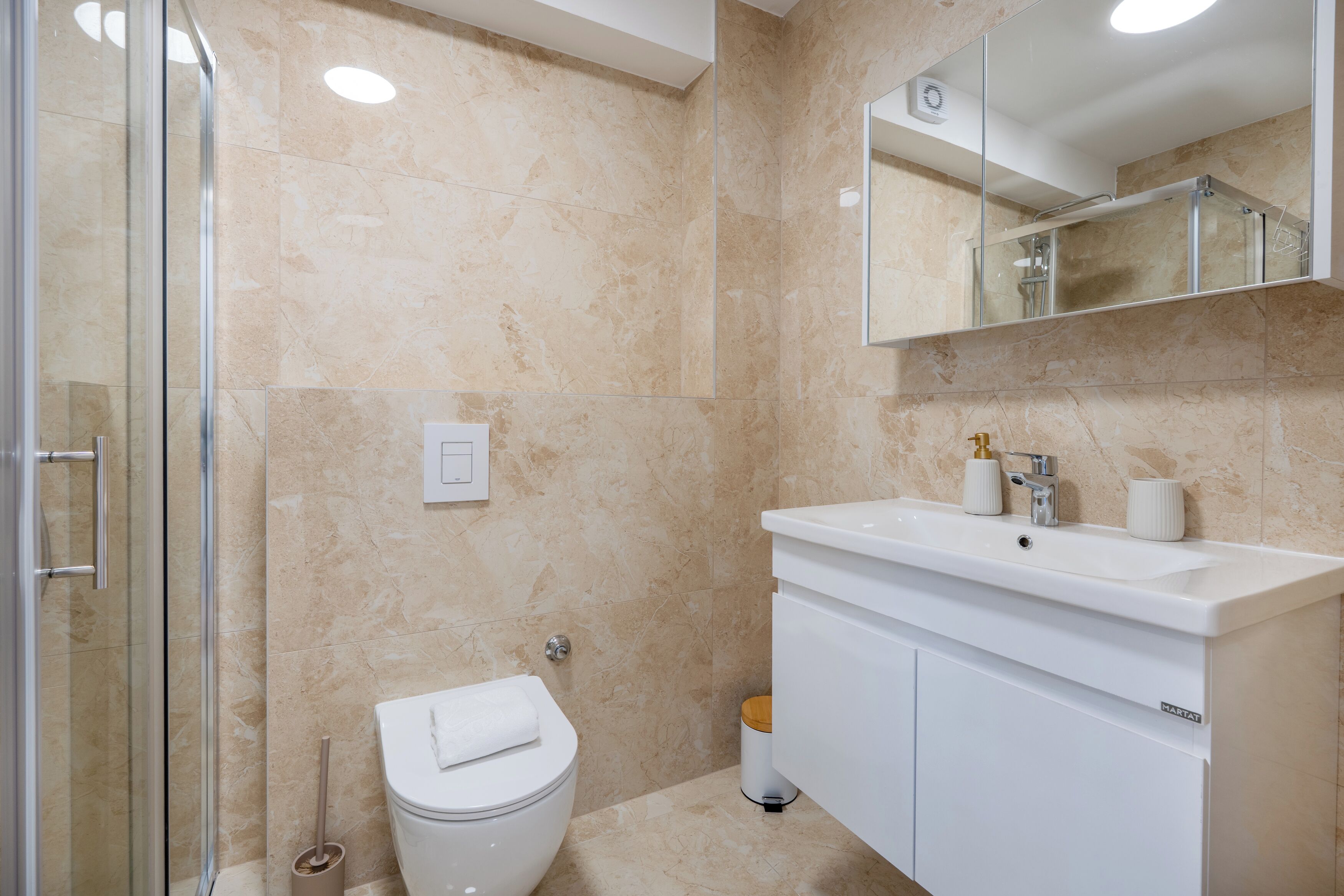 A bathroom with a glass shower enclosure, a white toilet, a sink with a mirror above it, and a trash bin.