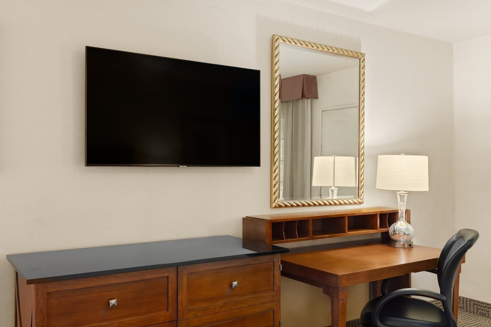 Una habitación de hotel con televisión de pantalla plana, un escritorio de madera con espejo, una lámpara y una silla.