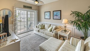 Un salon avec un canapé à rayures, un téléviseur, un ventilateur de plafond et une grande fenêtre donnant sur l’extérieur.