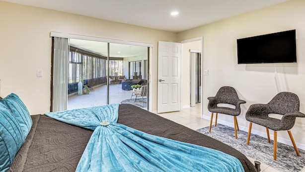 Un dormitorio con cama, televisor, dos sillas y puerta corredera que da a un balcón.